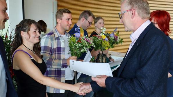 Person im Anzug überreicht Urkunde und schüttelt Hand einer Frau in schwarzem Top, weitere Personen mit Blumensträußen im Hintergrund.