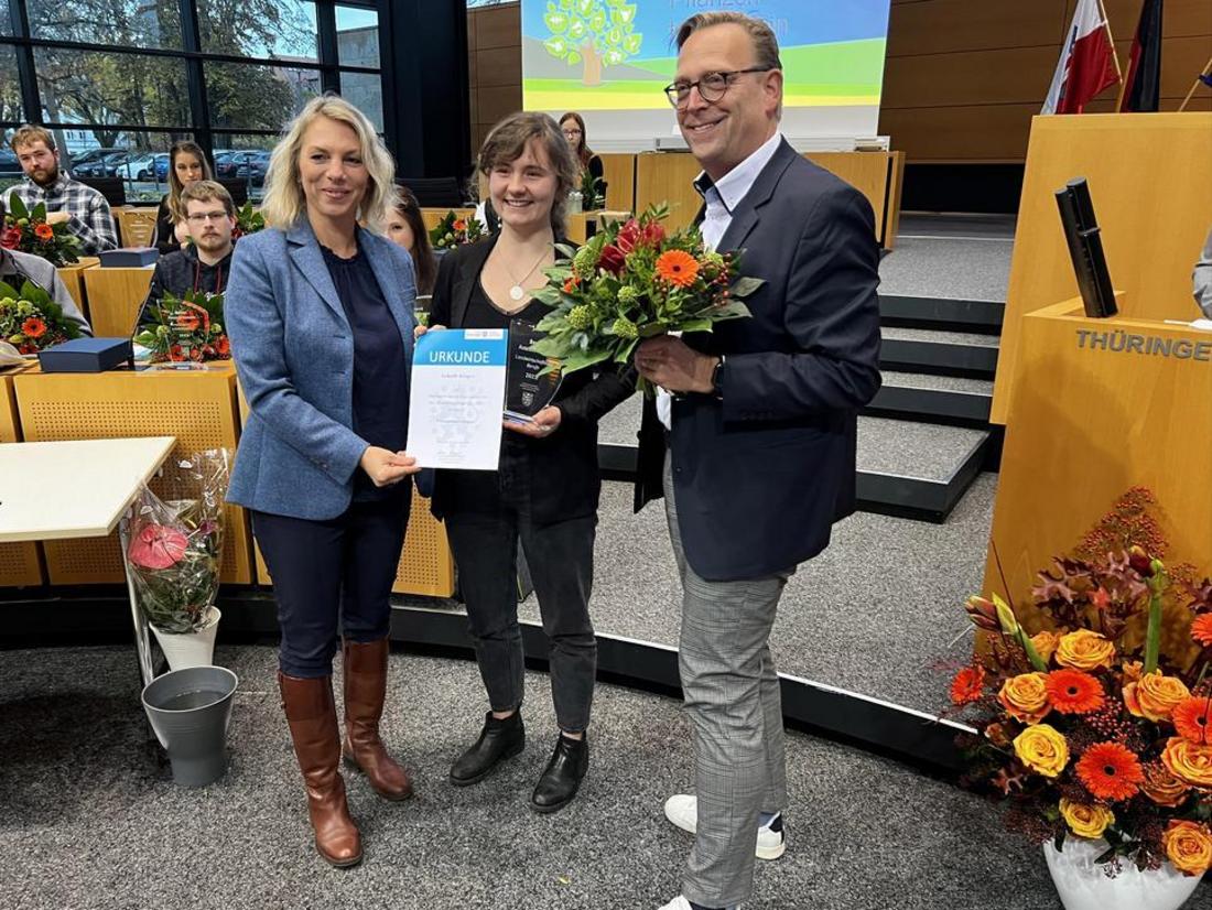 Das Bild zeigt eine Preisverleihung oder Ehrung. Drei Personen stehen im Vordergrund: zwei Frauen und ein Mann. Die Frau links hält eine Urkunde, die Frau in der Mitte hält einen Blumenstrauß, ebenso wie der Mann rechts. Im Hintergrund sind weitere Personen zu sehen, die im Publikum sitzen. Die Szenerie deutet auf eine formelle Veranstaltung hin. Im Hintergrund ist der Schriftzug "Thüringen" erkennbar.