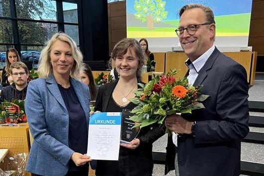 Das Bild zeigt eine Preisverleihung oder Ehrung. Drei Personen stehen im Vordergrund: zwei Frauen und ein Mann. Die Frau links hält eine Urkunde, die Frau in der Mitte hält einen Blumenstrauß, ebenso wie der Mann rechts. Im Hintergrund sind weitere Personen zu sehen, die im Publikum sitzen. Die Szenerie deutet auf eine formelle Veranstaltung hin. Im Hintergrund ist der Schriftzug "Thüringen" erkennbar.