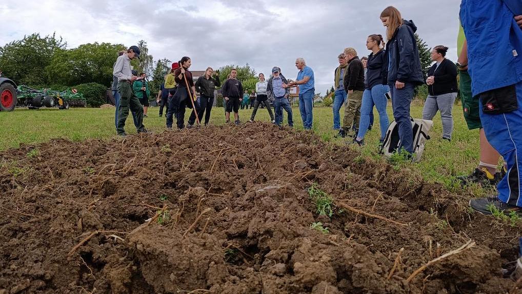 Das Bild zeigt eine Luftaufnahme eines Feldes, auf dem ein grüner Traktor mit einer blauen Saatmaschine arbeitet. Im Hintergrund sind einige Häuser und weitere landwirtschaftliche Maschinen zu sehen. Eine Gruppe von Menschen steht am Rande des Feldes und beobachtet die Arbeiten. Das Bild vermittelt den Eindruck einer Demonstration oder eines Feldtages für landwirtschaftliche Technik. Die Bearbeitung des Bodens wird dargestellt, möglicherweise zur Vorbereitung der Aussaat.