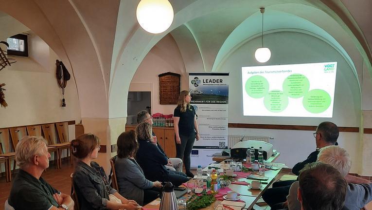 Eine Person steht vor einer Präsentation in einem Raum mit Bänken und Tischen. Auf dem Bildschirm sind grüne Punkte mit Text zu sehen. Die Anwesenden sitzen an einem langen Tisch, umgeben von Getränken und Notizen.