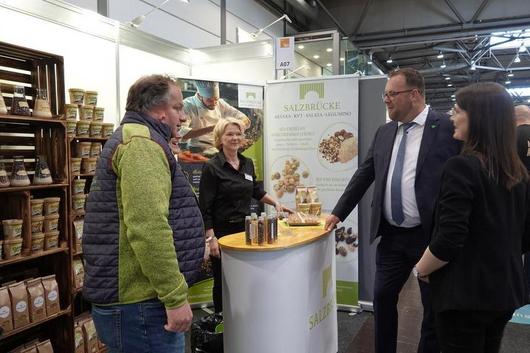 Vier Personen stehen an einem Messestand mit einem runden weißen Tresen und einem Banner mit der Aufschrift 'SALZBRÜCKE'