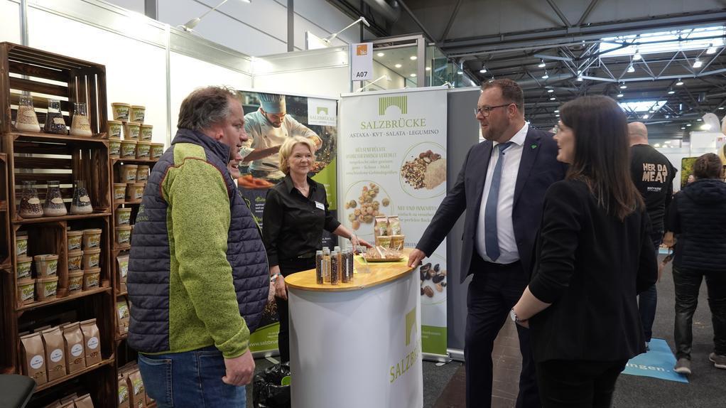 Vier Personen stehen an einem Messestand mit einem runden weißen Tresen und einem Banner mit der Aufschrift 'SALZBRÜCKE'