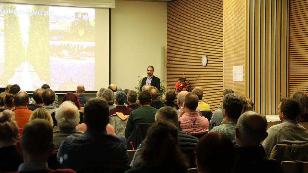 Vortragender steht vor einer Leinwand mit Präsentation vor einem Publikum in einem Raum mit Holzvertäfelung und gestreiften Vorhängen