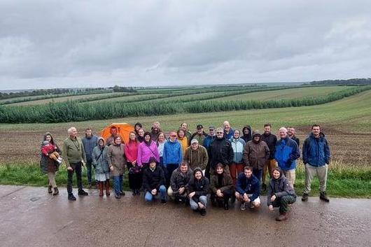 Gruppe von 27 Personen, teils stehend, teils kniend, auf einem nassen Weg vor Feldern unter bewölktem Himmel.