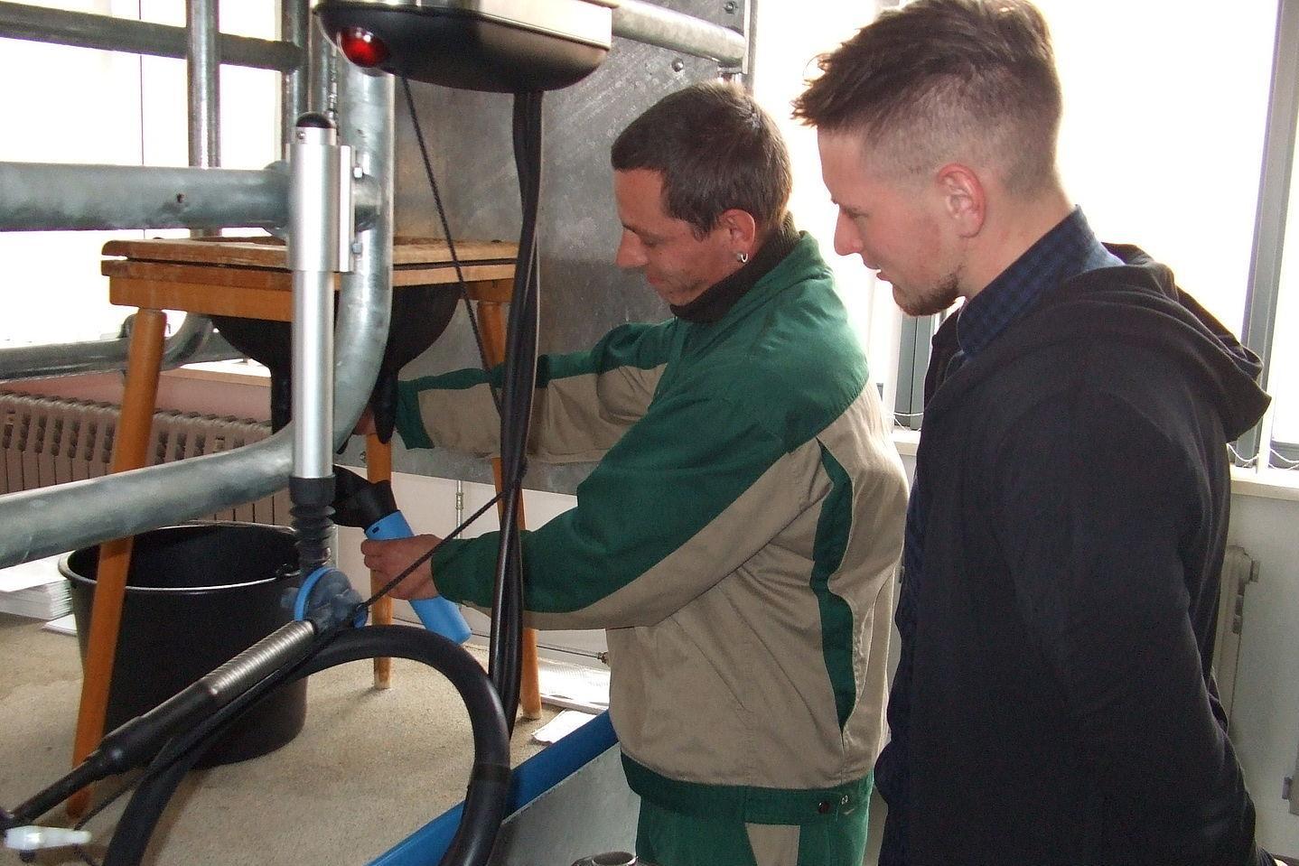 Zwei Männer in Arbeitskleidung bedienen eine Maschine mit Metallrohren in einem Innenraum.