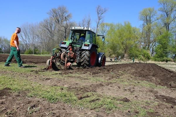 Traktor beim Pflügen mit Landarbeiter im Hintergrund, blauer Himmel und grüne Bäume.