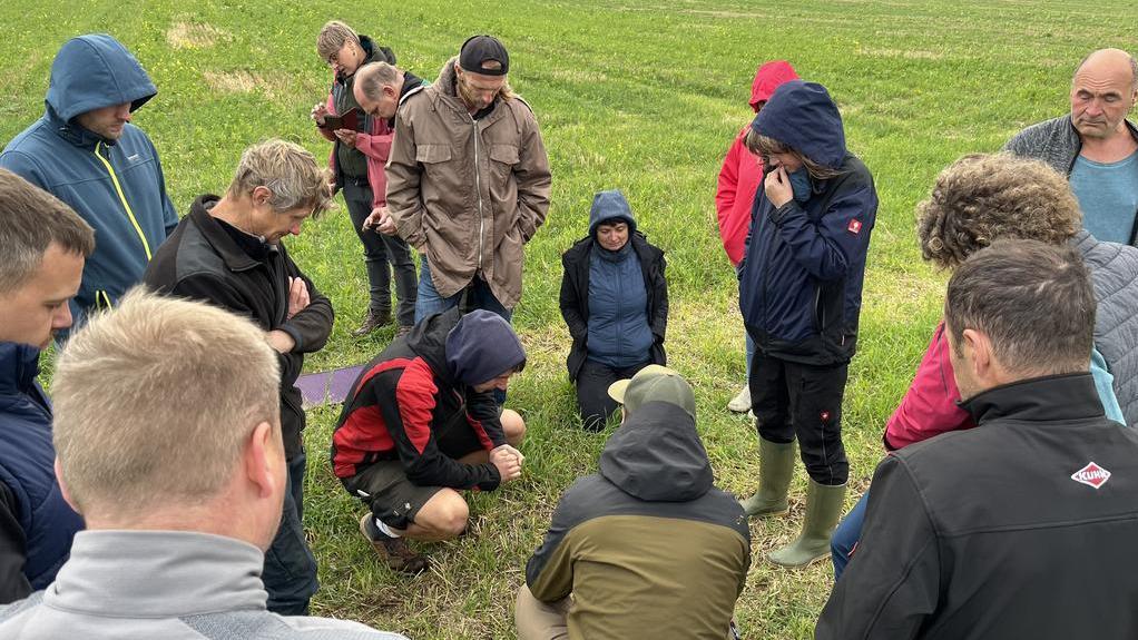 Gruppe von Menschen in Outdoor-Kleidung versammelt auf einem Feld, einige knien oder stehen, Blick auf den Boden gerichtet.