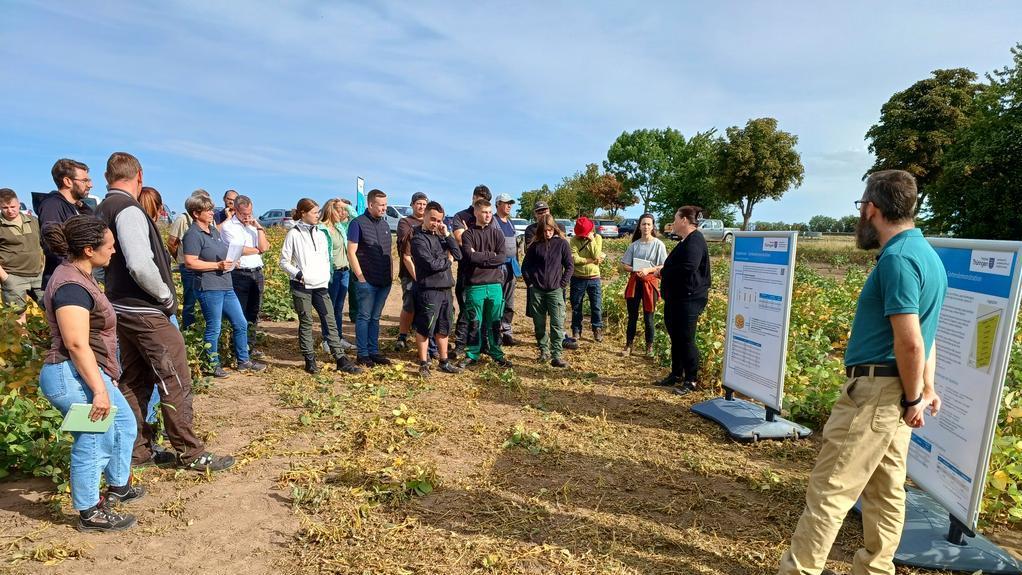 Das Bild zeigt eine Gruppe von Menschen, die auf einem Feld stehen und einem Mann zuhören, der vor zwei Plakaten steht. Der Mann hält einen Vortrag über Soja-Anbau, da die Pflanzen auf dem Feld wachsen.