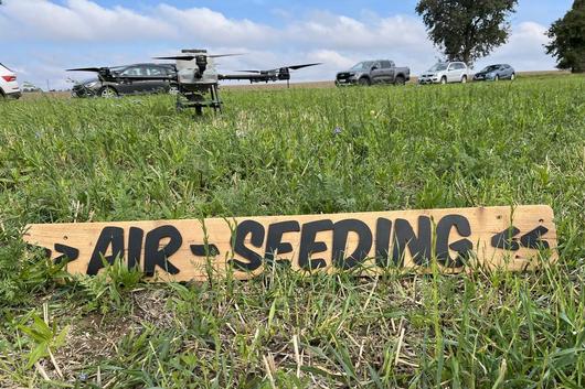 Drohne mit vier Rotoren steht auf einer Wiese hinter einem Holzschild mit der Aufschrift 'AIR SEEDING', im Hintergrund mehrere geparkte Autos und ein Baum.