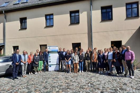 Gruppenfoto von 30 Personen vor einem Gebäude, einige tragen Anzüge, andere legere Kleidung. Im Hintergrund steht ein Banner mit dem Text 'LEADER'.