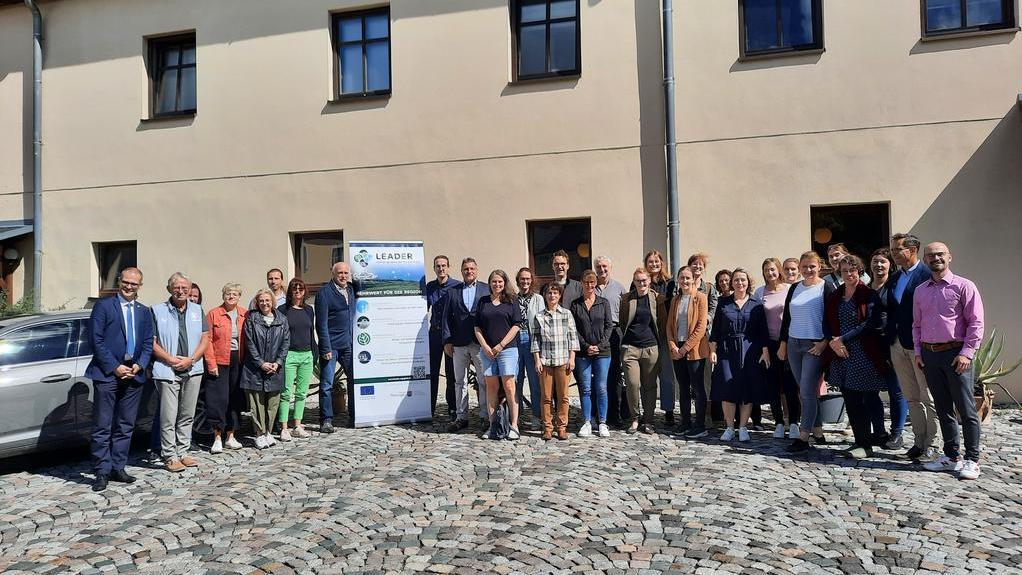 Gruppenfoto von 30 Personen vor einem Gebäude, einige tragen Anzüge, andere legere Kleidung. Im Hintergrund steht ein Banner mit dem Text 'LEADER'.