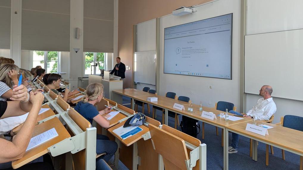 Ein Seminarraum mit einem Vortragenden an einem Pult. Auf einem großen Bildschirm wird eine Webseite angezeigt. Teilnehmer sitzen an Tischen und schreiben.